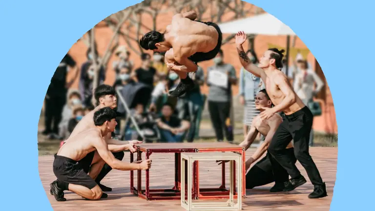 Five shirtless circus performers in an outdoor plaza balance and move red and white cube frames as one performer flips through the air above them, while a crowd watches in the background.