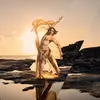 A dancer stands on a rock shelf in front of the ocean. It is sunrise and they are wearing a light yellow fabric that is flowing through the air.