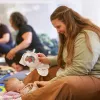 A woman sits smiling on a colorful play mat, engaging with a baby. In the background, other adults and babies interact, creating a warm, joyful scene.