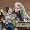 Two actors are seen on the set of the National Theatre Live production The Playboy of the Western World. A man sits leaning back on a desk while a woman leans over him from behind the desk.