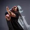 A dancer with long black hair, wearing a black t-shirt is looking upwards with their eyes closed. They are moving their arms in front of them.