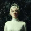 Marion Cotillard stands in a white outfit, with blonde hair and a silver crown. Snow is falling around her with a black backdrop.