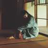 Japanese musician Ichiko Aoba sits on a wooden floor wearing a long faded green garment over the top of a red one that can only be seen near her feet and hands.