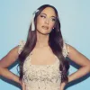 A white woman wears a blue bow in her long brown hair posing with her hands on her hips.