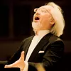 A man with white hairs and in black suit singing on the stage.