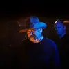 Mark Atkins is in a cowboy hat is lit with dramatic blue and orange lighting against a dark background. Erkki Veltheim stands blurred behind him, creating a mysterious tone.