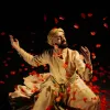 A man in traditional Persian attire gracefully dances amidst swirling red rose petals against a dark background.