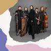 Five female musicians in black attire stand confidently against a gray backdrop. They hold various instruments, including a bass clarinet, global string instruments, and cello.