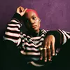 Arlo Parks wearing a striped knitted sweater, sitting on the ground against a dark purple wall.