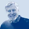 A white man with white hair and a beard smiling. He wears a blue sweater.