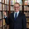 A man is standing in front of shelves of books. He is wearing glasses, a suit and tie and is holding up a pipe in his right hand.