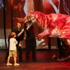 A young boy and two adults interact with a lifelike red dinosaur puppet on stage.