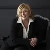 A woman with blonde hair sits in a regal looking black chair. She is wearing a black blazer and trousers and a white shirt with a wide collar.