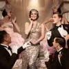 A glamorous 1920s-style scene shows a woman in a sparkling gown on a staircase, surrounded by dancers in pink costumes and tuxedos striking theatrical poses.