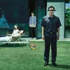 A promotional image from Parasite showing a family standing and lounging outside a modern glass-walled home, each with black bars obscuring their eyes. One man stands in the foreground while another holds a large stone near the doorway. The scene has an e