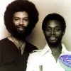Vintage black-and-white portrait of two smiling men, one with a large afro and beard and the other with short hair and a collared shirt.