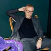 A white man in a casual grey suit and glasses sits relaxed in a chair.