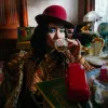 A woman in a red hat and plaid coat with bright blue eyeshadow drinks from a glass, surrounded by eclectic, vintage decor and a retro telephone.