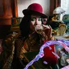 A woman in a red hat and plaid coat with bright blue eyeshadow drinks from a glass, surrounded by eclectic, vintage decor and a retro telephone.