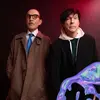 The two members of sparks stand facing the camera in front of a red background.