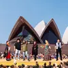 Three men and three women from different backgrounds stand on a yellow platform in front of the Sydney Opera House sails in front of an audience.