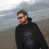 A man stands on a beach wearing a hoodie and sunglasses. He is looking off to his right.
