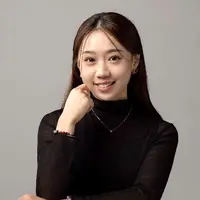 A woman with long black hair is holding her hand to her cheek and smiling. She is wearing a black shirt and silver necklace.