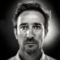 A man looks straight on, he is wearing a white collared shirt and has stubble. His hair is dark and sticking up.