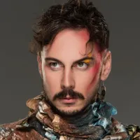 Man with brown hair and moustache stares powerfully to the side, and has red streaked makeup on one side of his face.