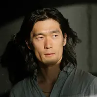 A man looks in to the distance with light in his eyes. He is wearing a collared shirt and has shoulder length black hair.