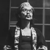 A black and white image of a man wearing a robe and glasses, with a grey streak in his shoulder length hair.