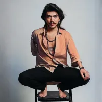 Man with long hair and a mustache crouches on a chair. He wears a partially open shirt with beads and black pants.