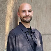 A man in a black tshirt and dark blue button shirt stands against a wall smiling.