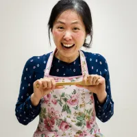 A woman with a big grin is holding a wooden spoon out in front of herself. She is wearing a blue shirt and floral apron.