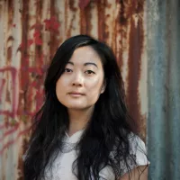 A Taiwanese, Australian woman with long hair standing in front of a rusty wall.