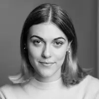A woman is seen from the shoulders up looking forward. She is wearing a white sweater and has shoulder length hair.