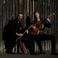 Two men are seating, one holds a guitar and one holds a flute-like instrument.