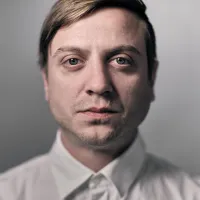 Composer Mischa Blanos is looking straight ahead wearing a white collared shirt in front of a white background.