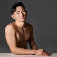 A shirtless man sits at a table. He is resting his arm on the table and looking off to his left.