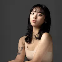 A woman in a beige singlet sits with her arms in front of her. A tattoo is visible on one arm.