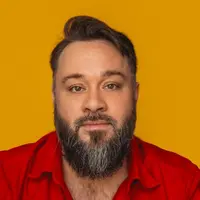 A man in a red collared shirt with short hair is standing against a yellow backdrop.