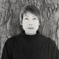 A woman with short hair wearing a black jumper is standing with her back against a tree.