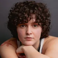 Person with short curly hair leans forward with their hand on their chin.