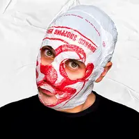 A man in front of a white textured background is wearing a black shirt and a plastic bag over his head. The white bag with red design has eye holes and a mouth hole cut out.