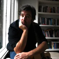 A man sits while resting his chin in his hand. Behind him is a shelf full of books.