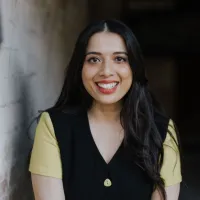 A woman with long black hair is smiling. She is wearing a black buttoned top with yellow sleeves.