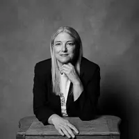 A woman with light straight hair sits with one hand under her chin and one resting on a wooden desk. She is wearing a white shirt with a blazer on top.