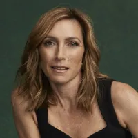 A women with brown hair is wearing a sleeveless black top looking straight on. Behind her is a dark green background.