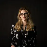 A woman standing against a black background. She has long light brown hair, glasses and a black shirt on.