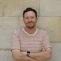 A man standing against a sandstone wall. He is wearing translucent glasses and wears a striped t-shirt.
